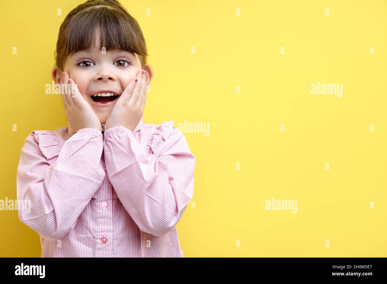 little child girl is surprised by something, holding hands on cheek. cute kid in pink blouse in shock, isolated on yellow studio background Stock Photo