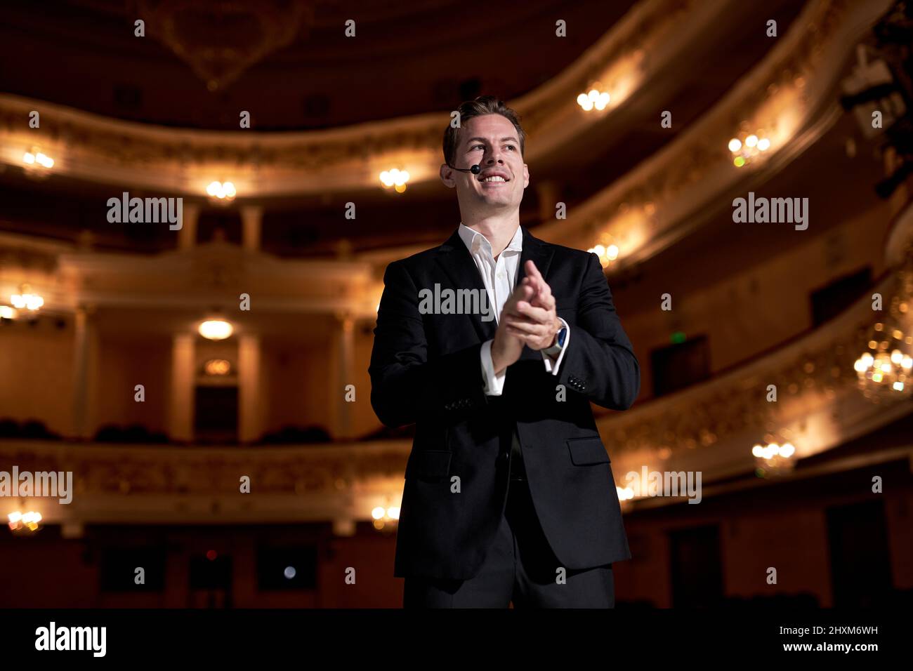 Confident guy of american appearance in formal classic suit clapping hands after performance, on stage. Presenter Speaking to Audience People. Unident Stock Photo