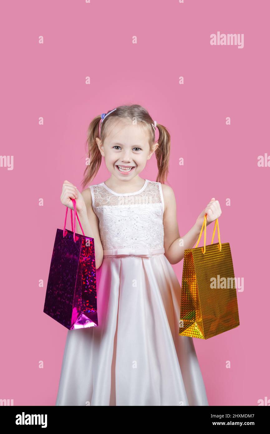 Cute little caucasian girl in a party dress on pink background happy enjoying shopping holding colorful bags Stock Photo