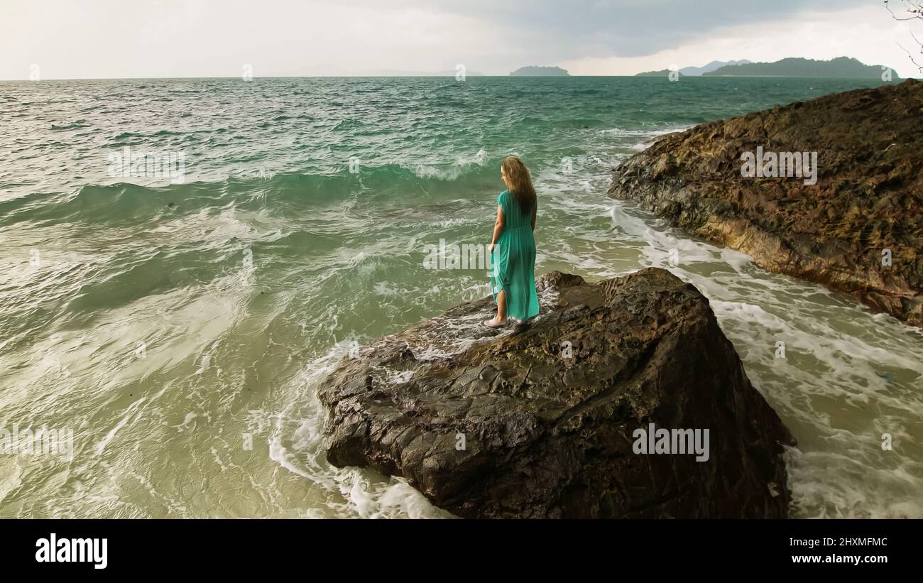 Seductive long-haired woman walks on rock of sea reef stone, stormy cloudy ocean. Woman in blue swimsuit dress tunic. Concept rest in sea, tropical re Stock Photo