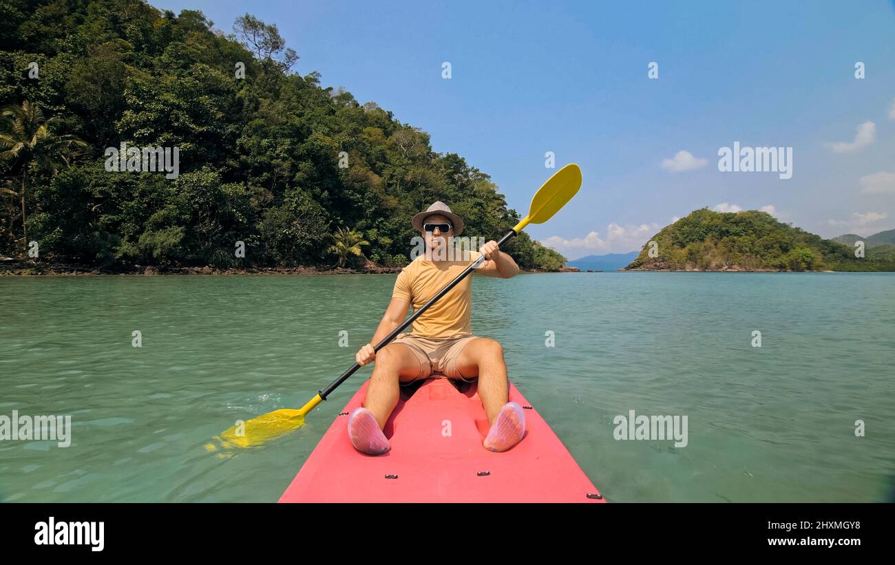 Young man with sunglasses and hat rows pink plastic canoe along sea against green hilly islands with wild jungles. Traveling to tropical countries. Strong guy is sailing on kayak in ocean, front view. Stock Photo
