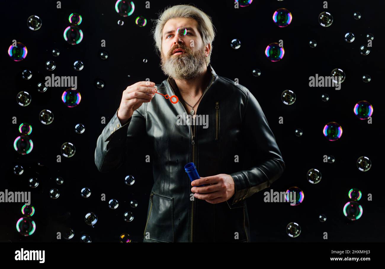 Bearded Man play with Soap bubbles. Happiness. Childhood. Guy in leather jacket blowing bubble. Stock Photo