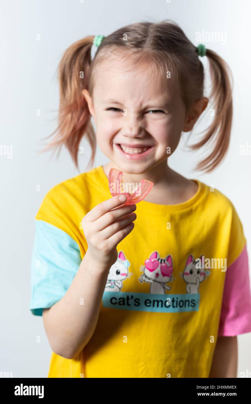 Cute little caucasian girl with blond hair is holding a pink dental myofunctional trainer on white the background. dental tariner is made to help equa Stock Photo