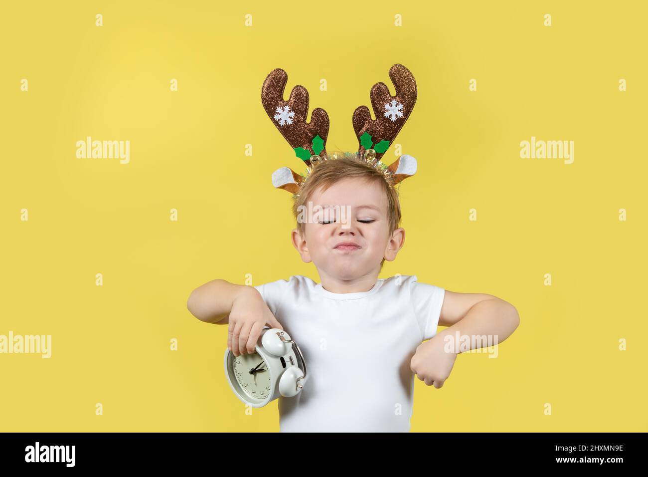 Cute caucasian boy in in christmas reindeer costume with alarm clocks in his hands making funny faces. A place for your text. Studio shot on a yellow Stock Photo