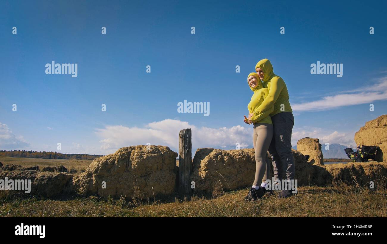 Man and woman in yellow green sportswear. Lovely couple of travelers hug and kiss near old stone enjoying highland landscape. Two travelers are walking against the backdrop of snow-capped mountains. Stock Photo