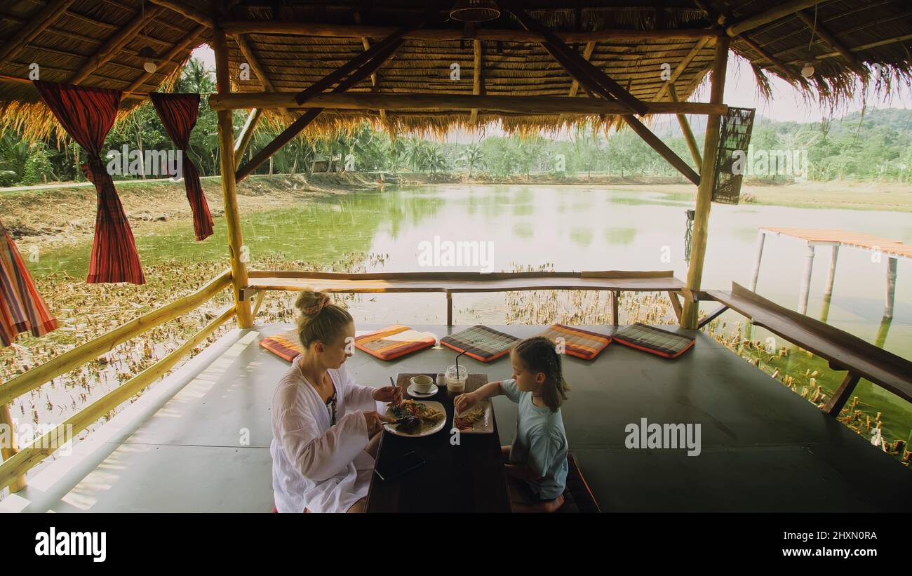 The happy family eat and drink tasty beverages spending time in local floating cafe on water. Mother and daughter having breakfast outdoors. Woman and a child in a cafe. Tropical view Stock Photo