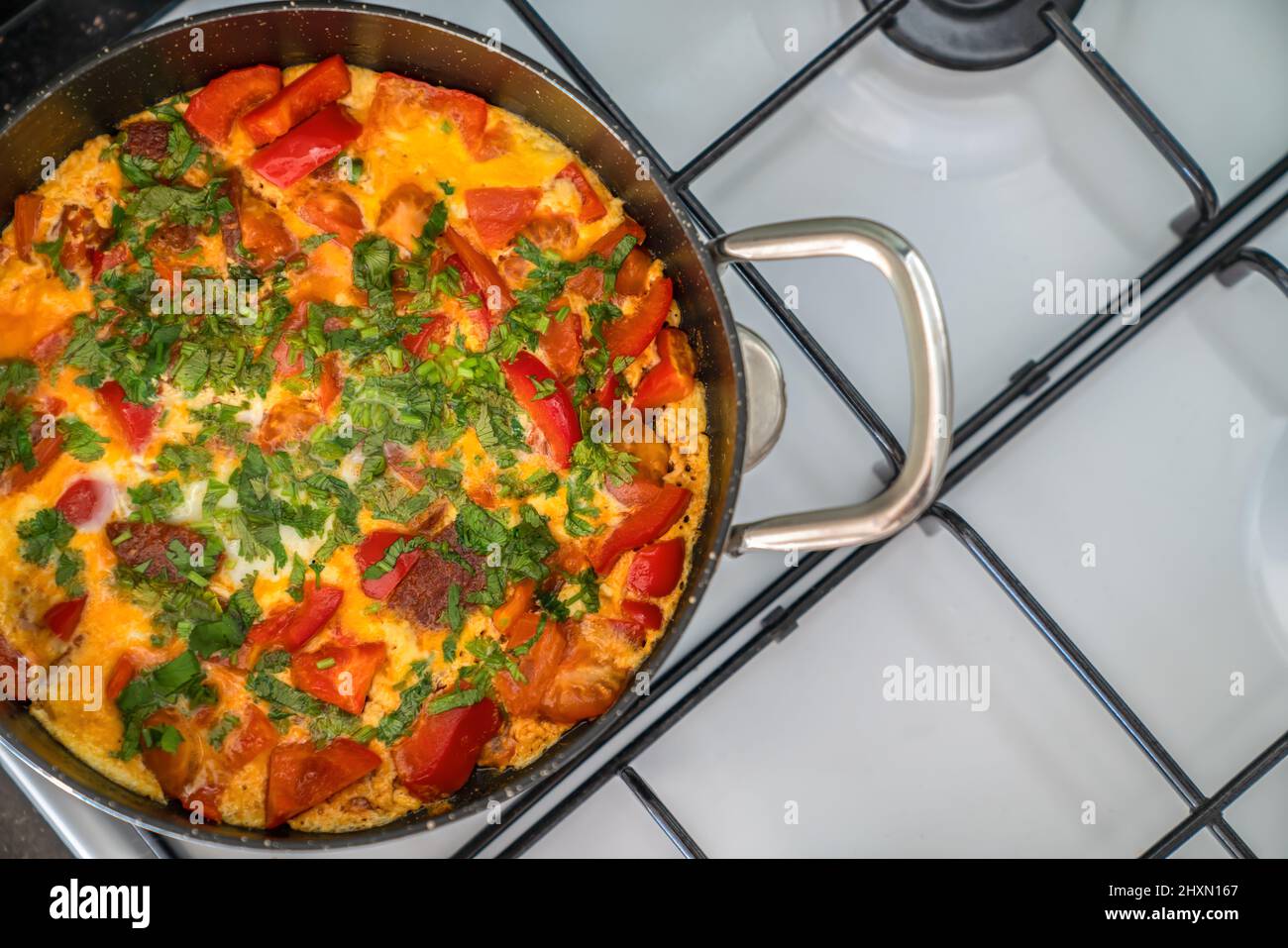 Omelette with vegetables and herbs in black frying pan on white stove. Top view Stock Photo