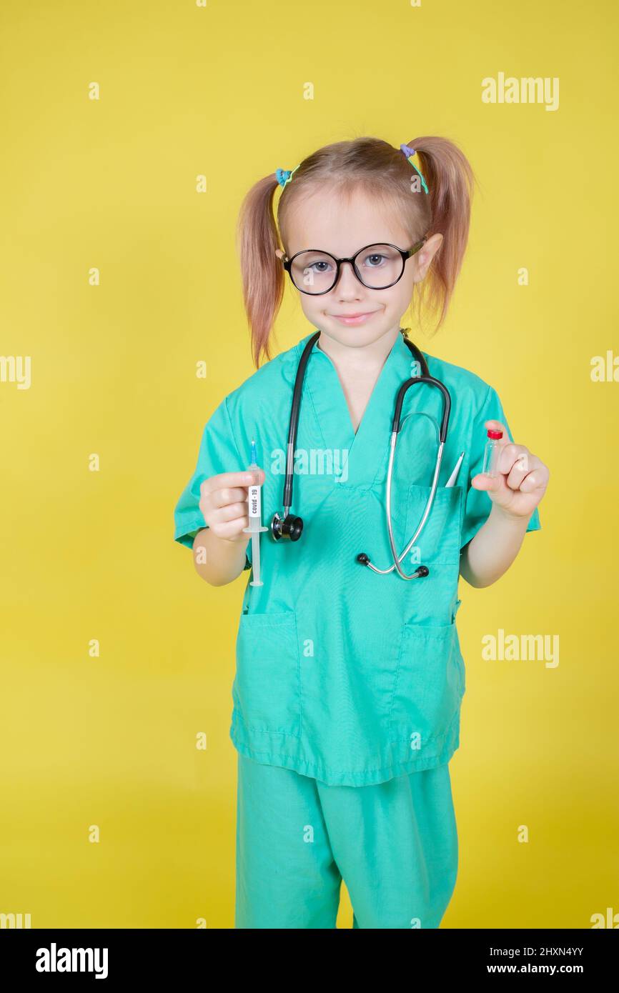 Portrait of little caucasian girl dressed in doctors green coat with syringe and medicine bottle in hand on yellow background Stock Photo