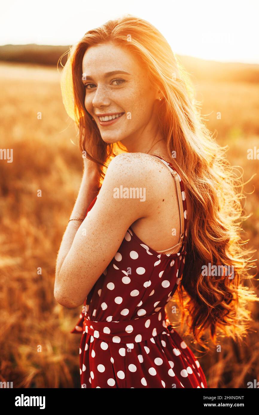 Portrait of a red-haired young lady with a freckled face looking and smiling at the camera while enjoying nature. Portrait on light backdrop. Wheat Stock Photo
