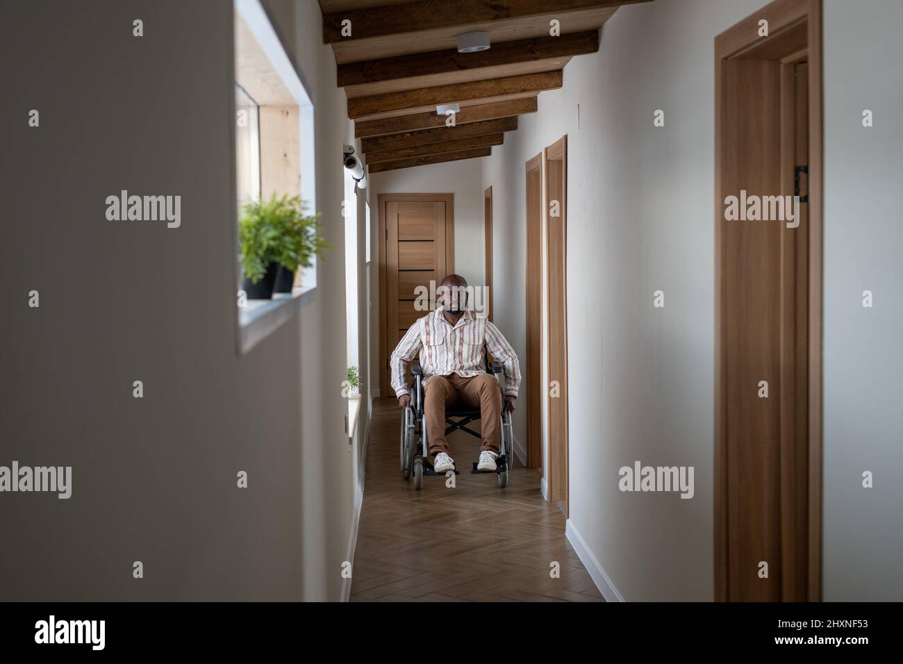Young man of African ethnicity with disability moving along corridor of contemporary apartment while sitting in wheelchair Stock Photo