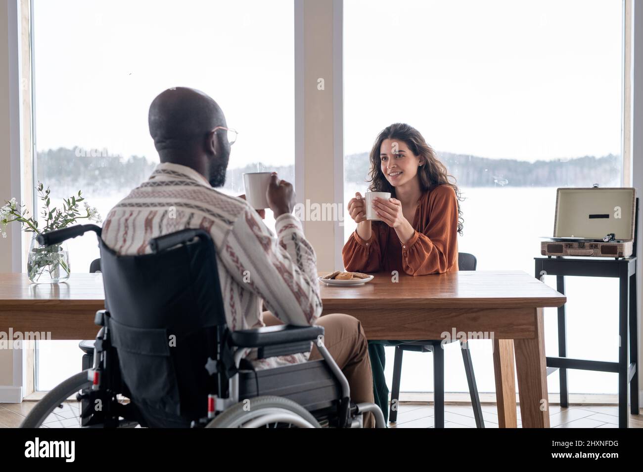 Happy young female with mug sitting by kitchen table and looking at black man with disability during conversation by breakfast Stock Photo