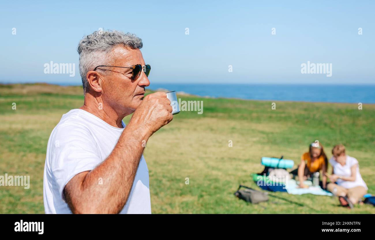 Senior man with sunglasses drinking coffee outdoors with his family in background Stock Photo