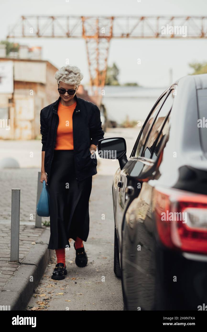 Stylish Young Woman in Jacket and Sunglasses Moving Near the Black Car Stock Photo