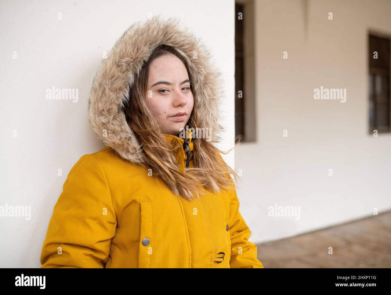 Thoughtful young woman with Down syndrome weraing parka, leaning the wall and looking at camera. Stock Photo