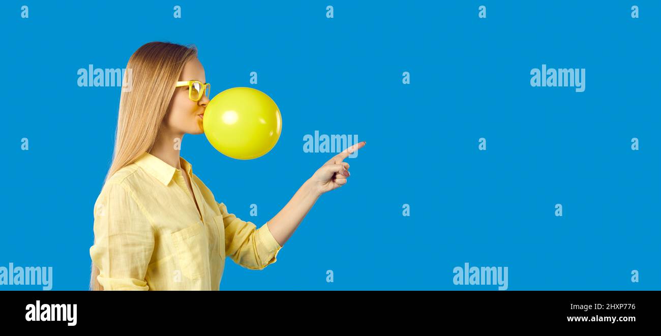 Young woman blowing up yellow balloon and pointing her finger at copy space on the right Stock Photo