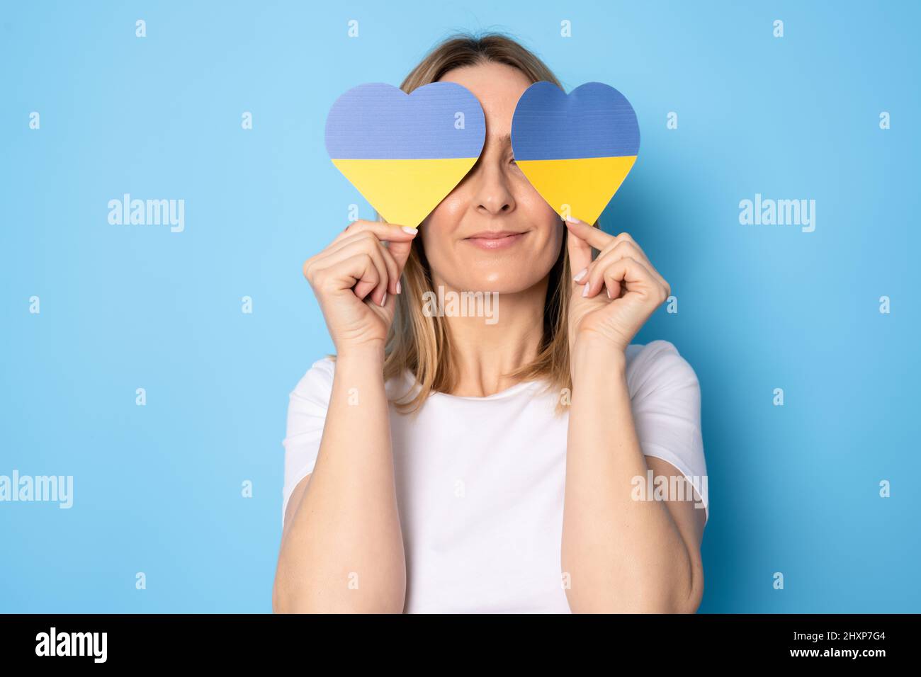Young smiling woman showing ukraine paper hearts standing isolated over blue background. Stock Photo