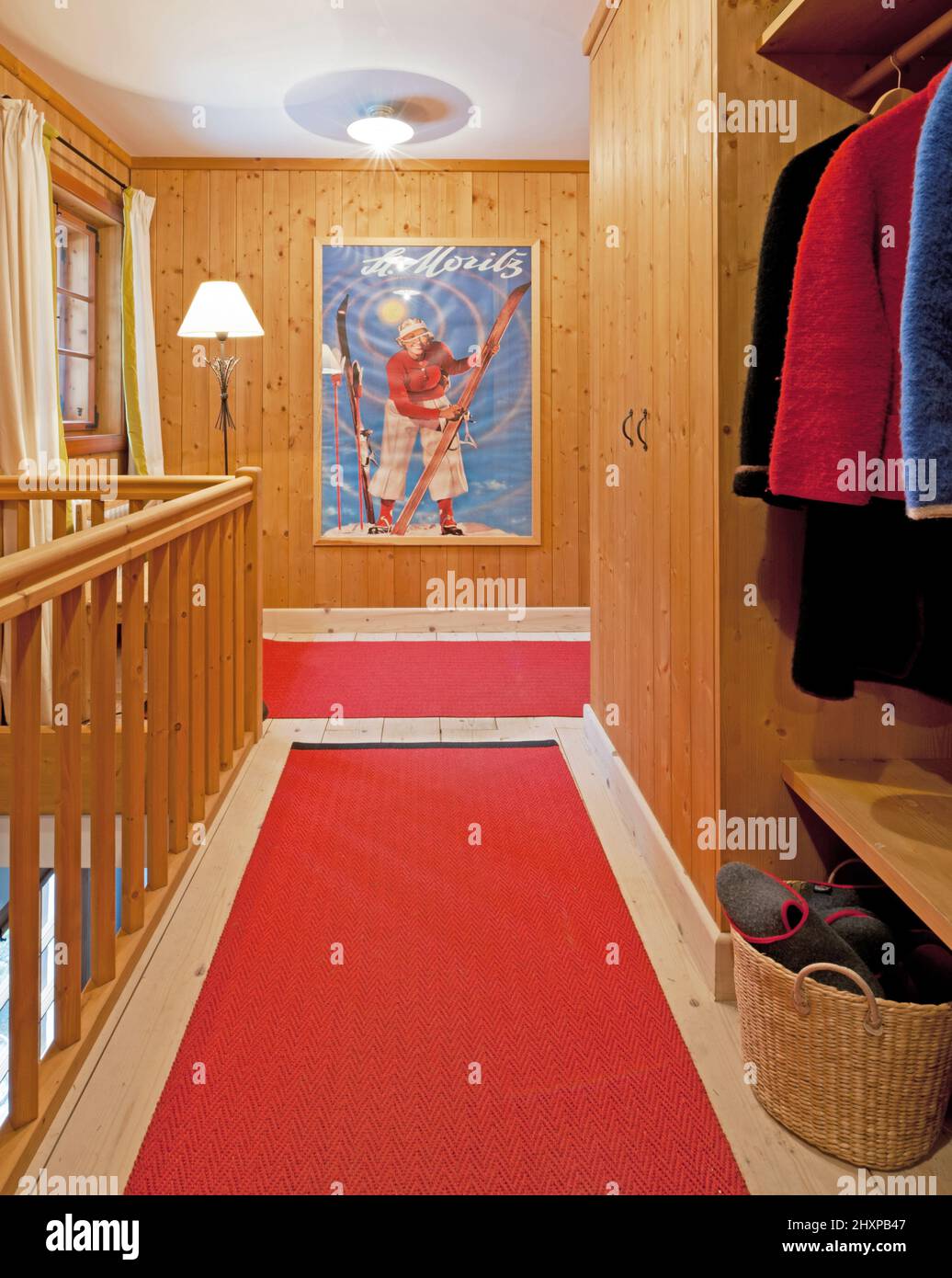 entrance hall in Alpine style house with wood panelled walls, wooden railings, red carpet, wardrobe with loden jackets, lamps and blue poster Stock Photo