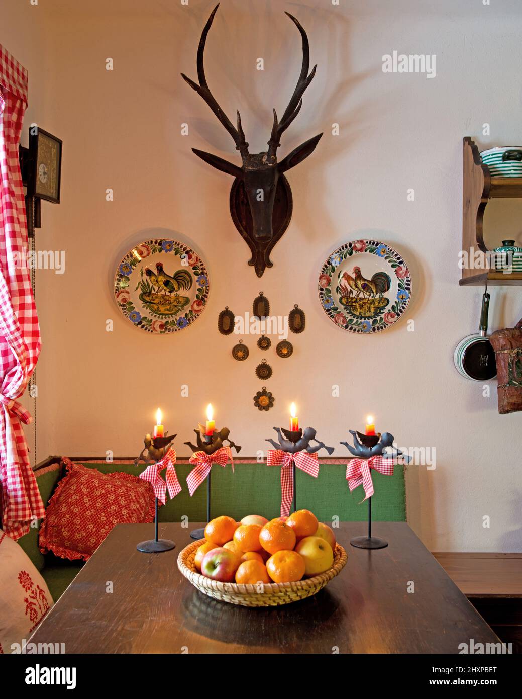country kitchen with table decorated with Christmas candles and basket with fruits, bench with cushions, antler and plated on the wall Stock Photo