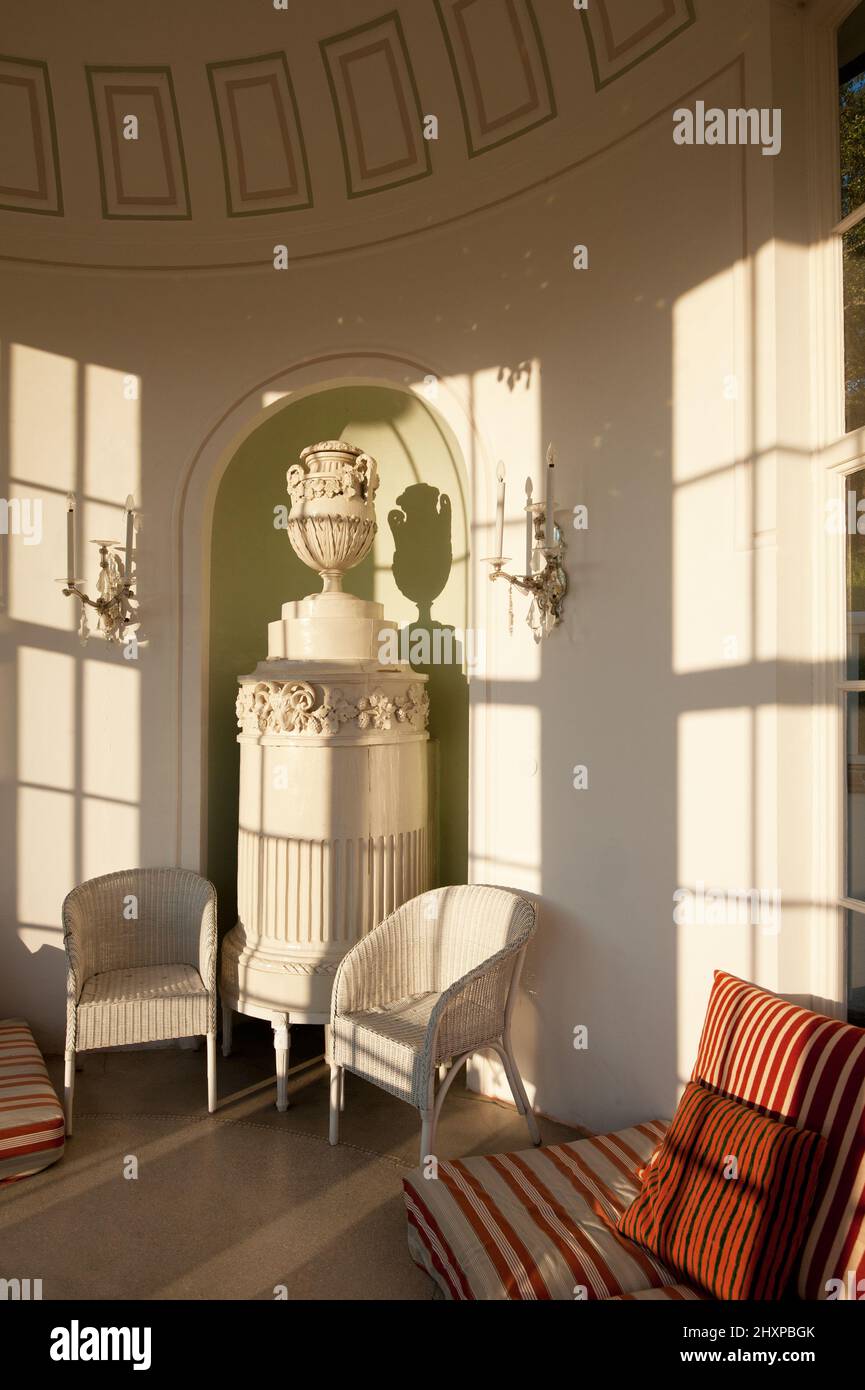 19th century garden pavillion with richly decorated tiled or ceramic stove, two white rattan chairs and striped cushionin lated afternoon sunlight Stock Photo