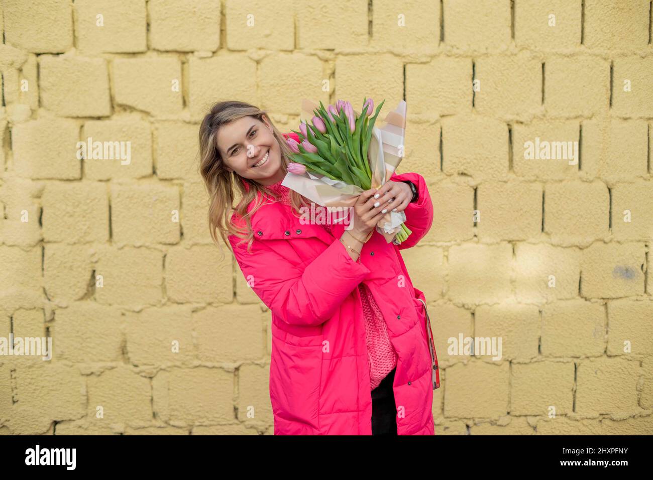 Glad embarrassed pretty woman holding present pink fresh flowers tulips, in pink jacket, looking at camera. Mother day Stock Photo
