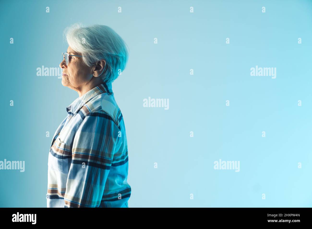Sad and pensive older gray-hared lady standing profile. Medium studio shot over blue background. High quality photo Stock Photo