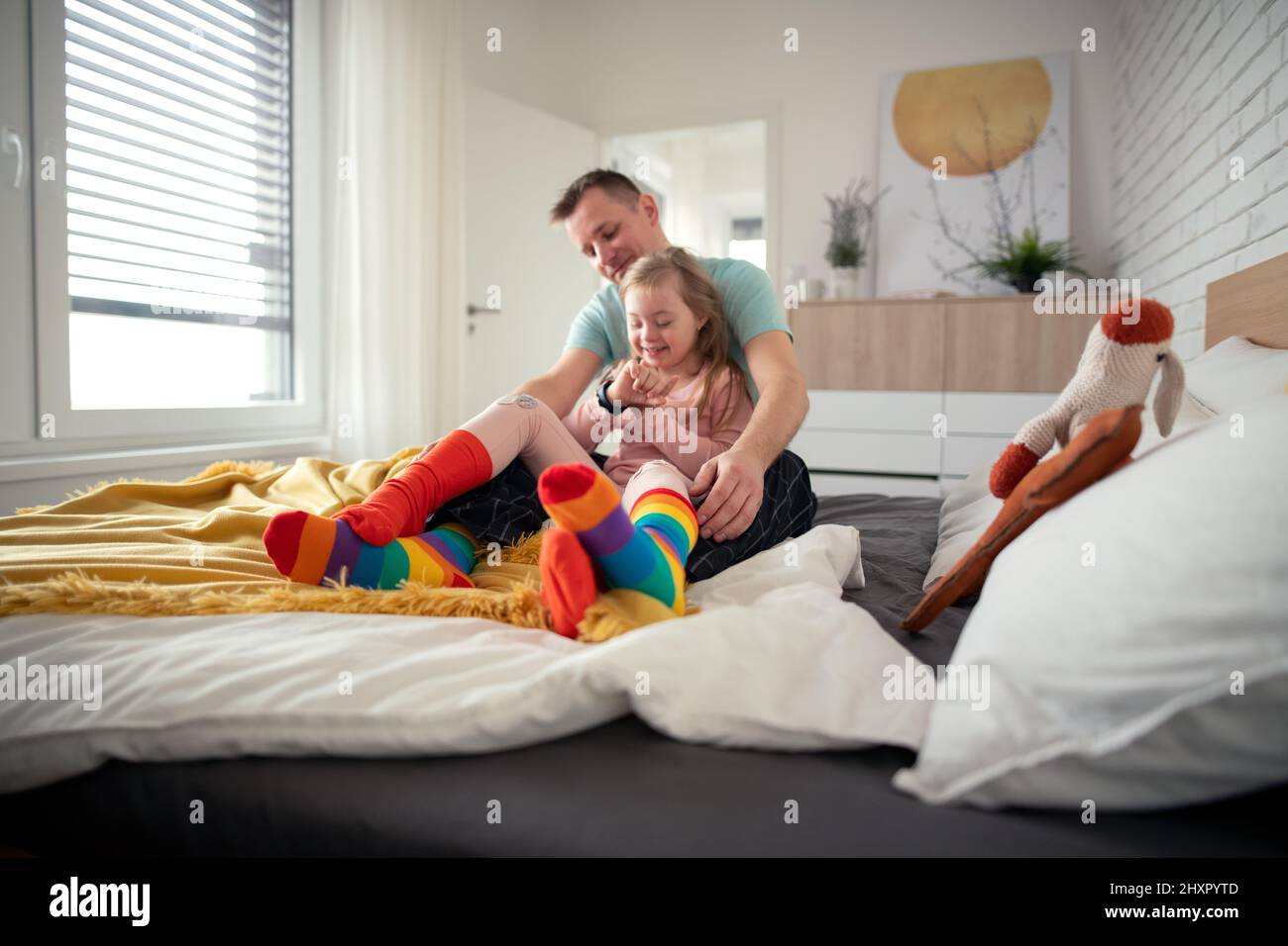 Father putting on different socks to his little daughter with Down syndrome when sitting on bed at home. Stock Photo