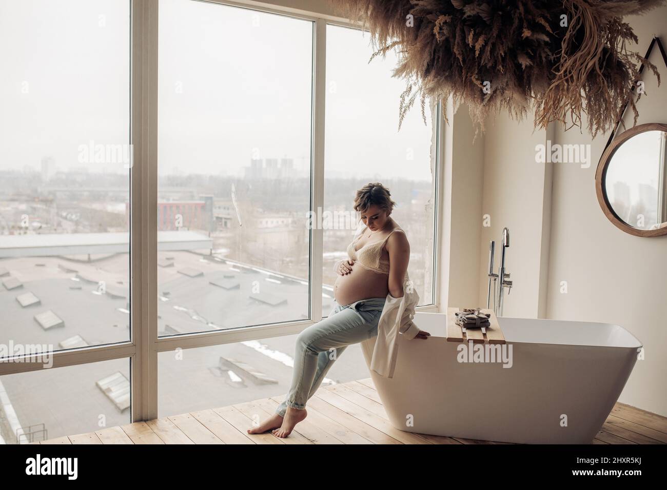 Young happy pregnant woman sitting on the edge of bathtub in bathroom against window background ...