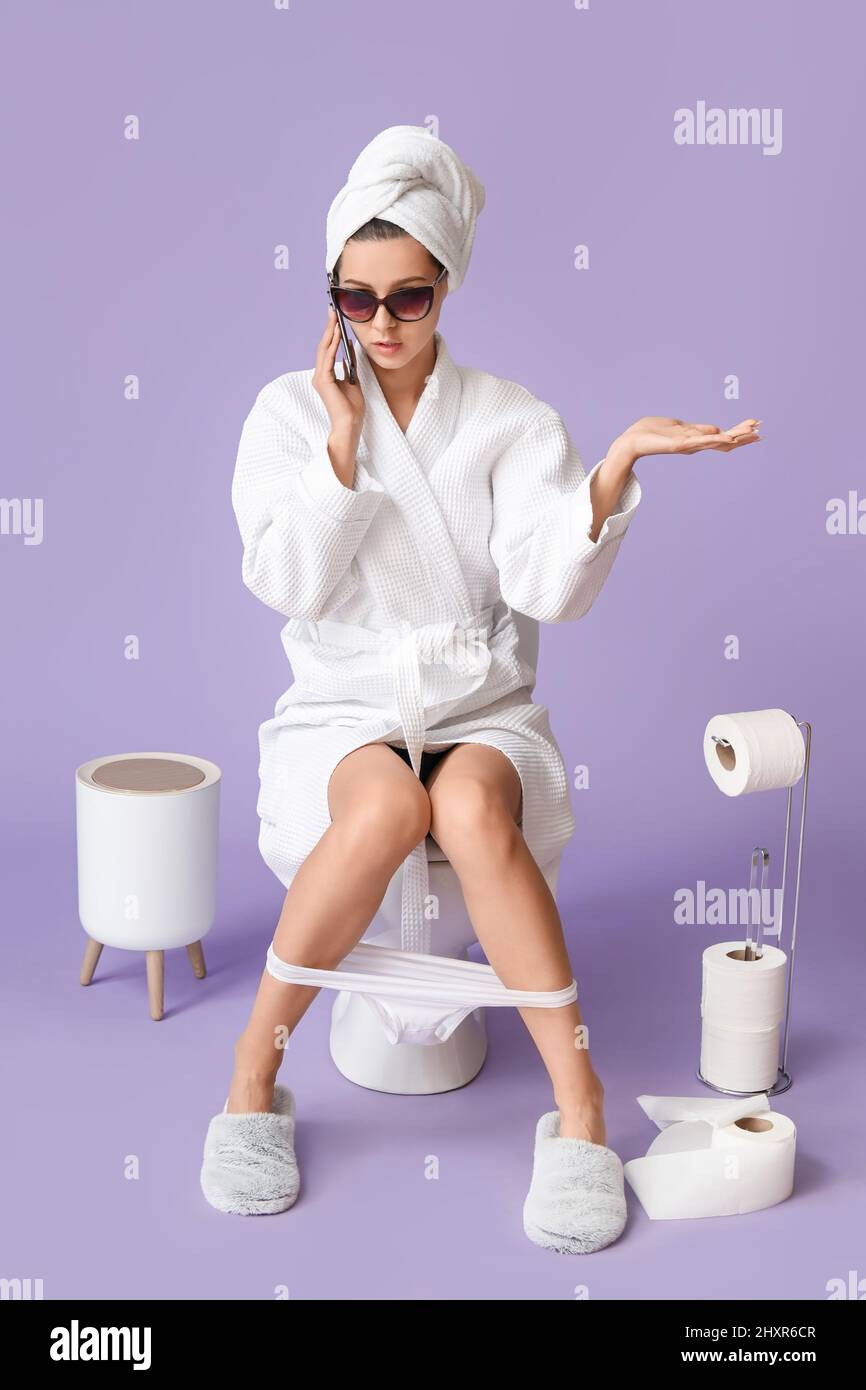 Cool young woman talking by phone while sitting on toilet bowl against