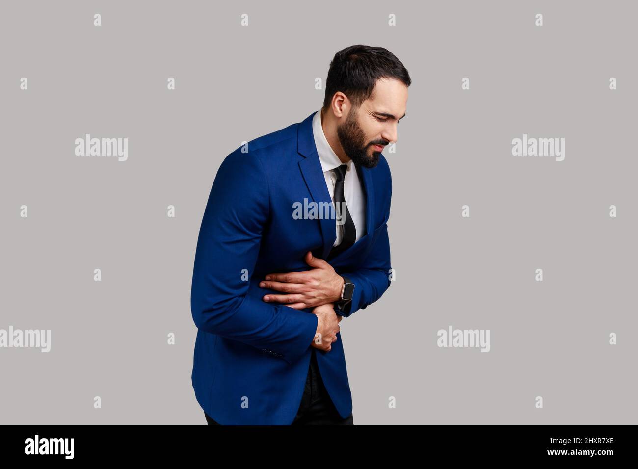 Bearded man touching belly, grimacing from stomach ache, severe abdominal distress, symptoms of constipation, indigestion, gastrointestinal disorder. Indoor studio shot isolated on gray background. Stock Photo