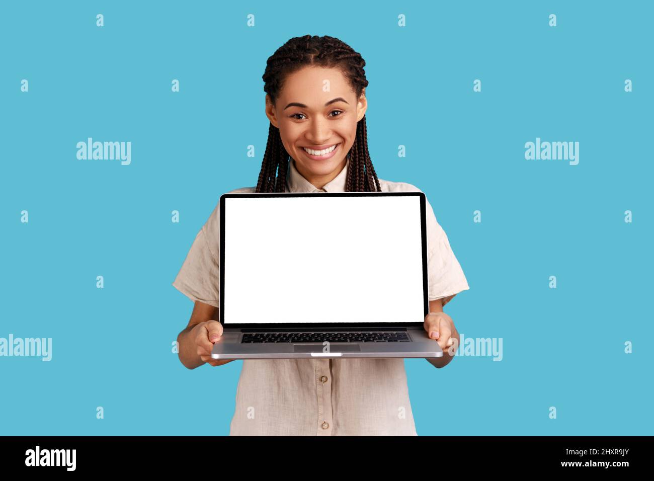 Happy woman with black dreadlocks shop assistant advertises new device, holding laptop with empty screen, expresses satisfaction, wearing white shirt. Indoor studio shot isolated on blue background. Stock Photo