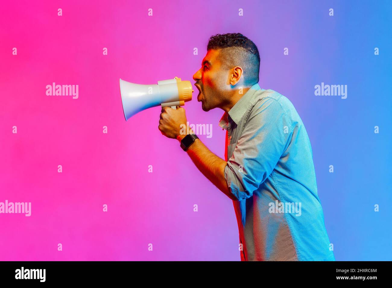 Side view portrait of angry man in shirt loudly speaking screaming holding megaphone, announcing important message. Indoor studio shot isolated on colorful neon light background. Stock Photo