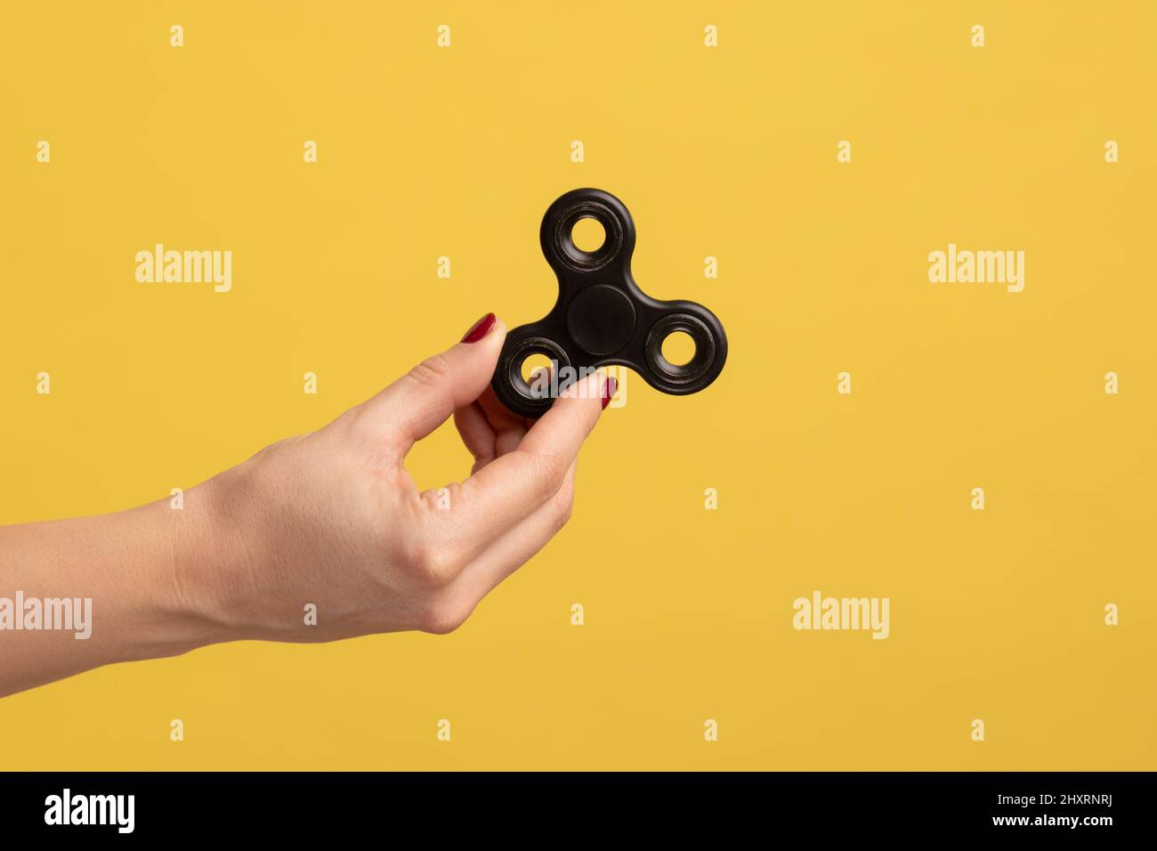 Closeup side view profile portrait of woman hand holding black fidget spinner, stress relieving toy. Indoor studio shot isolated on yellow background. Stock Photo