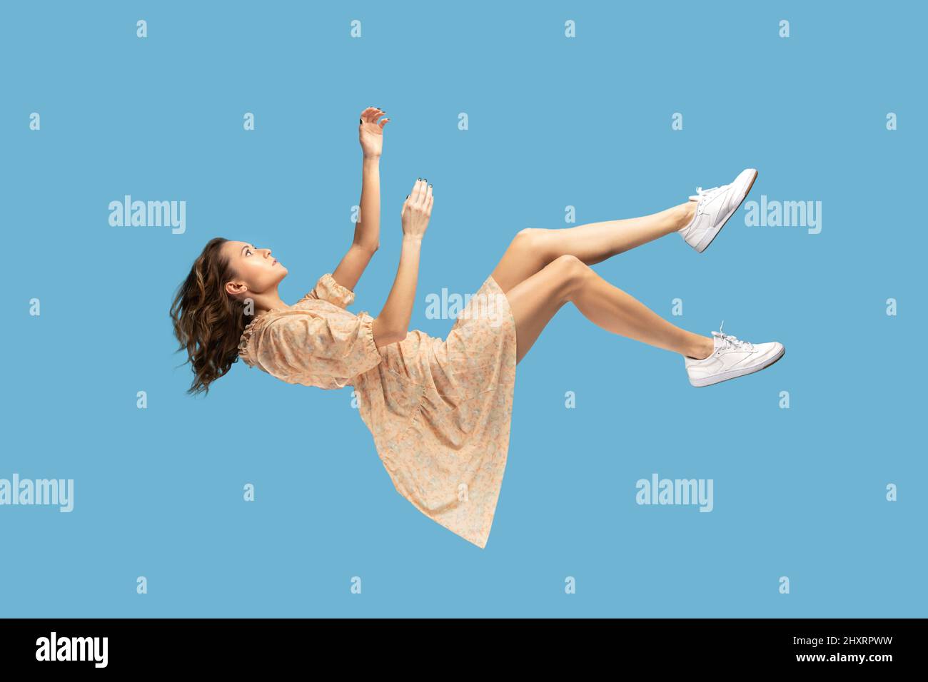 Floating in air. Relaxed girl in yellow dress levitating, looking up while flying mid-air, having comfortable peaceful dream. full length studio shot isolated on blue background, indoor Stock Photo