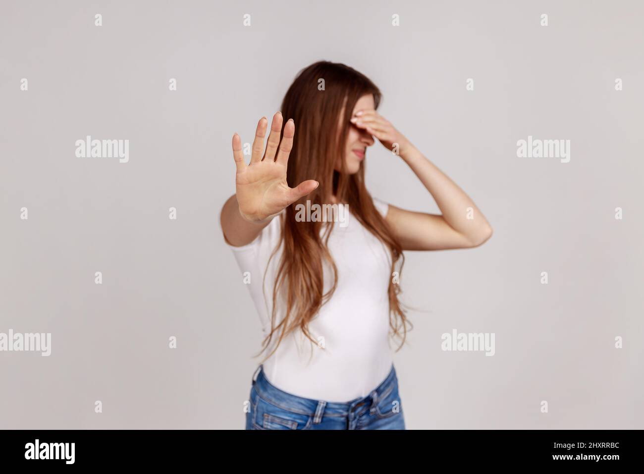 Portrait of afraid beautiful woman closing eyes with palm and showing stop hand gesture, turning face, does not want to see, wearing white T-shirt. Indoor studio shot isolated on gray background. Stock Photo