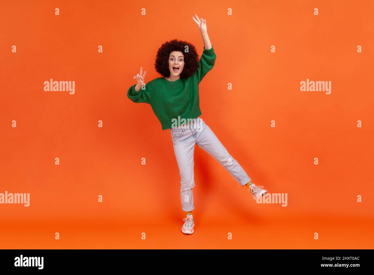 Full length portrait of amazed happy woman with Afro hairstyle wearing green casual style sweater standing on one leg, showing victory gesture, v sign. Indoor studio shot isolated on orange background Stock Photo