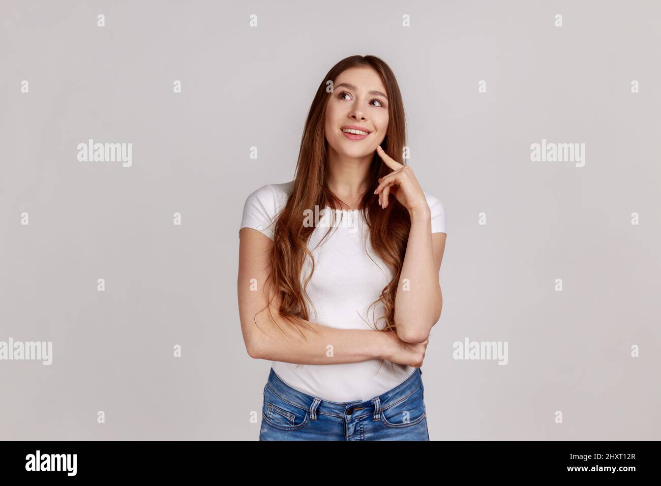 Portrait of adorable dreamy woman standing looking away, keeping finger on cheek, dreaming, making wish, love, wearing white T-shirt. Indoor studio shot isolated on gray background. Stock Photo
