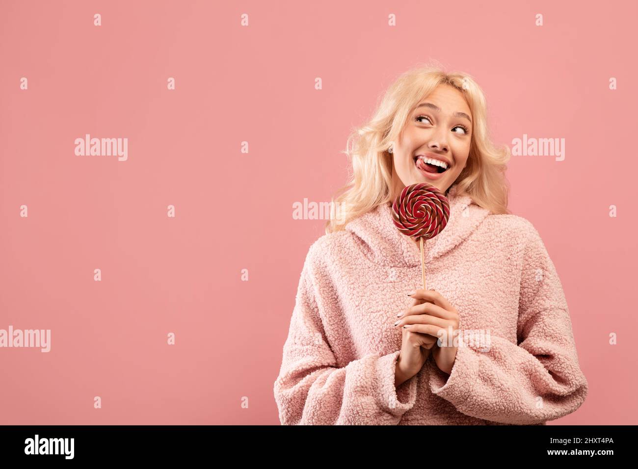 Sweet tooth concept. Playful lady holding candy on stick, looking aside at free space and smiling, pink background Stock Photo