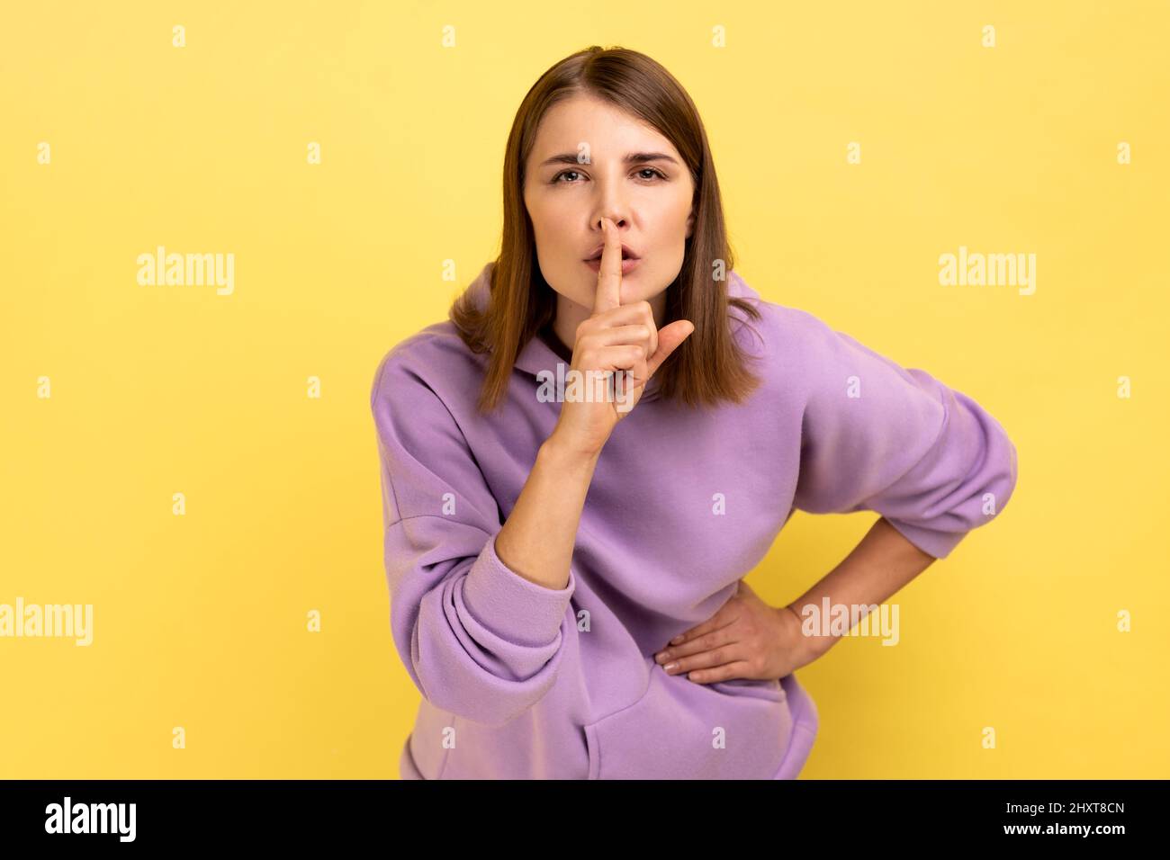 Shh, it's big secret. Portrait of beautiful serious woman showing gesture secret sign with finger near her lips, wearing purple hoodie. Indoor studio shot isolated on yellow background. Stock Photo