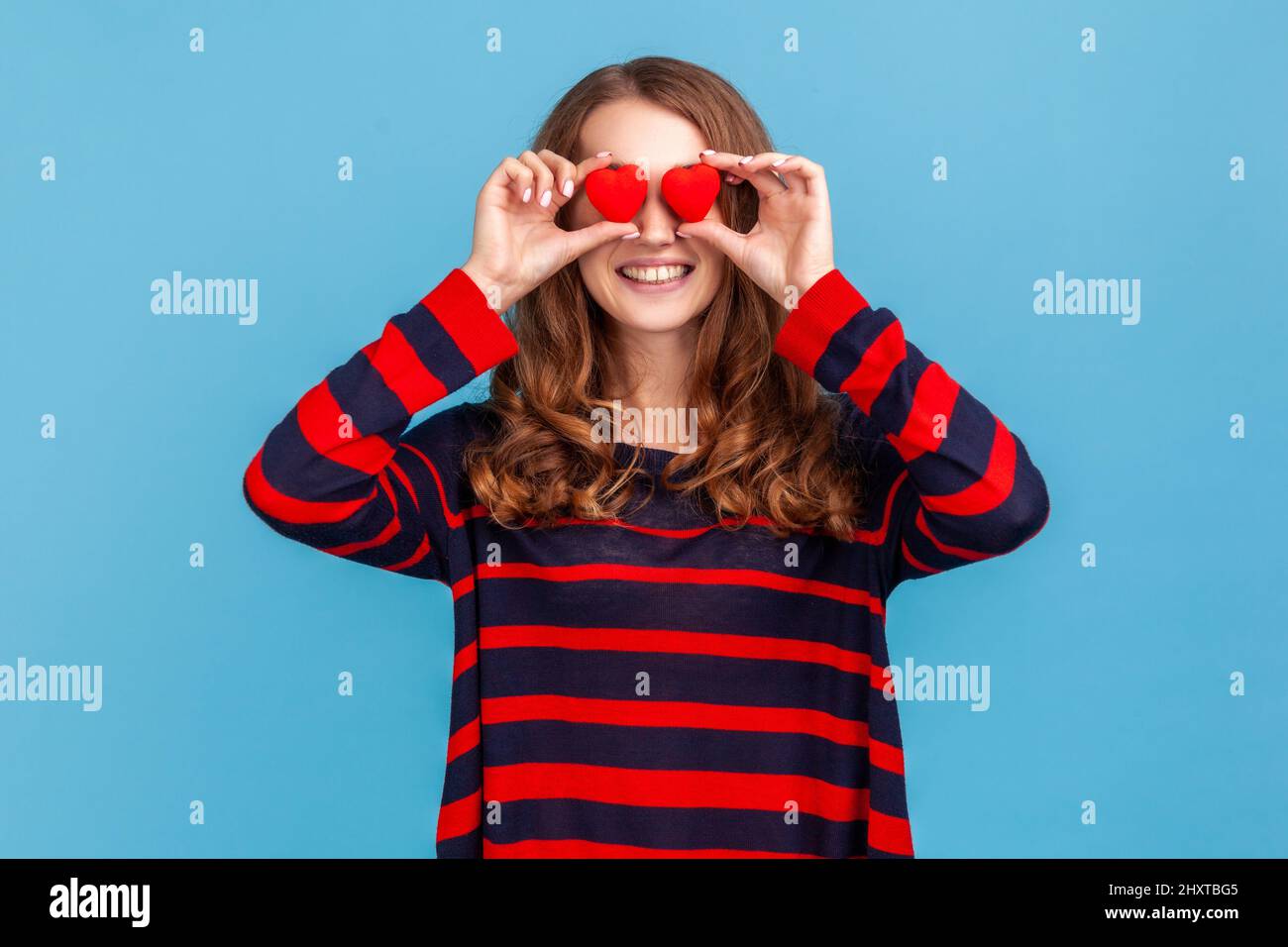 Funny positive woman wearing striped casual style sweater, covering eyes with small red hearts, love symbol, smiling toothily, falling in love. Indoor studio shot isolated on blue background. Stock Photo