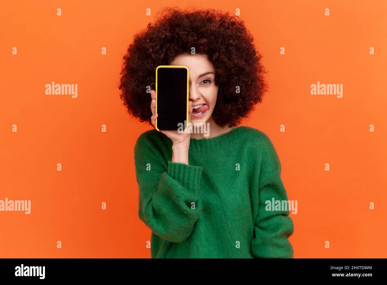 Positive funny woman with Afro hairstyle wearing green casual style sweater covering eye with cell phone with blank screen and showing tongue out. Indoor studio shot isolated on orange background. Stock Photo