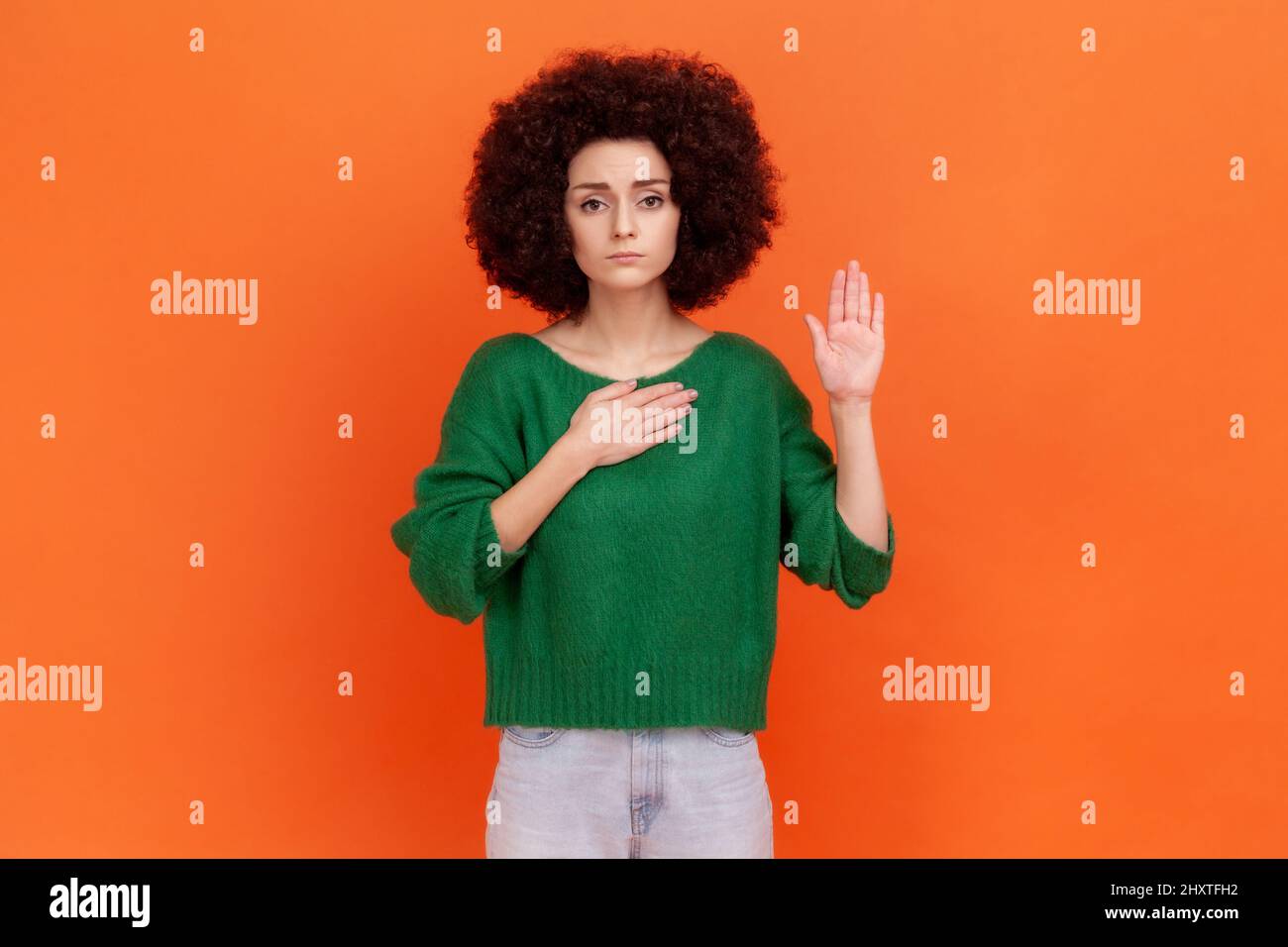 I promise. Serious woman with Afro hairstyle wearing green sweater standing raising hand and saying swear, making loyalty oath, pledging allegiance. Indoor studio shot isolated on orange background. Stock Photo