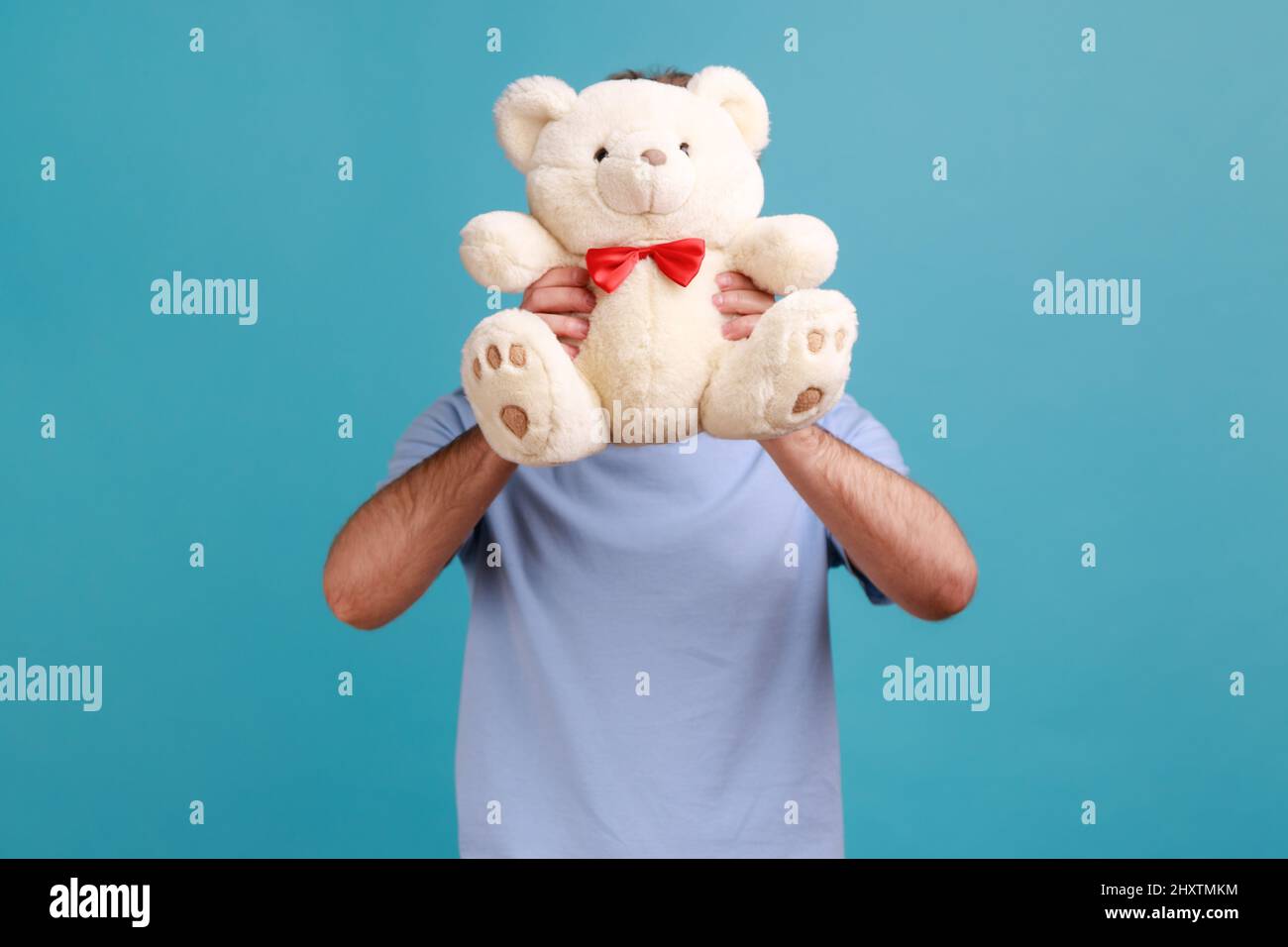 Portrait of unknown anonymous bearded man hiding his face behind white soft teddy bear, romantic surprise for his girflriend. Indoor studio shot isolated on blue background. Stock Photo