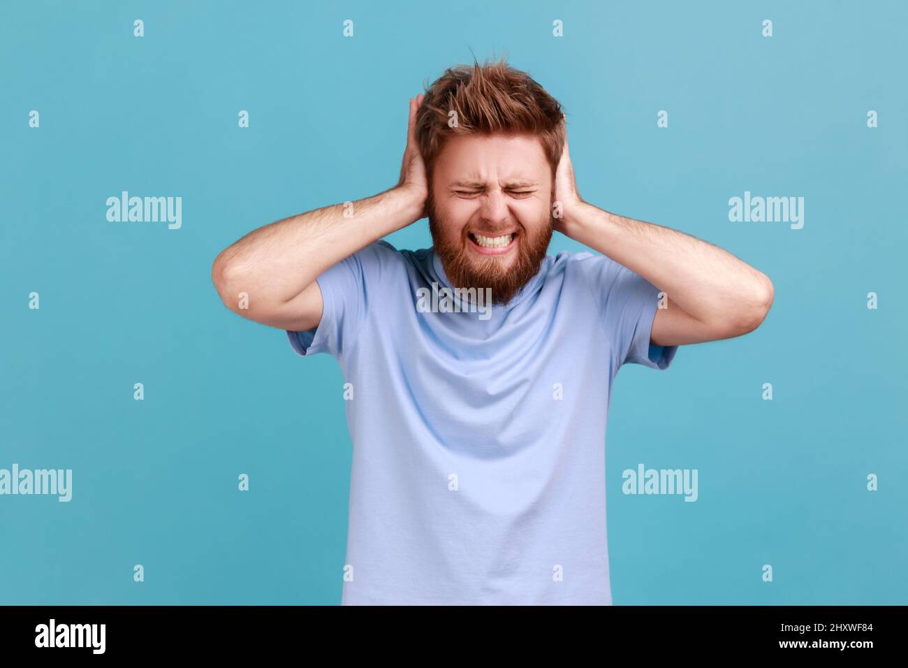 Don't want to listen. Portrait of despaired man irritated by loud noise covering ears and grimacing in pain, suffering annoying high-decibel sound. Indoor studio shot isolated on blue background. Stock Photo