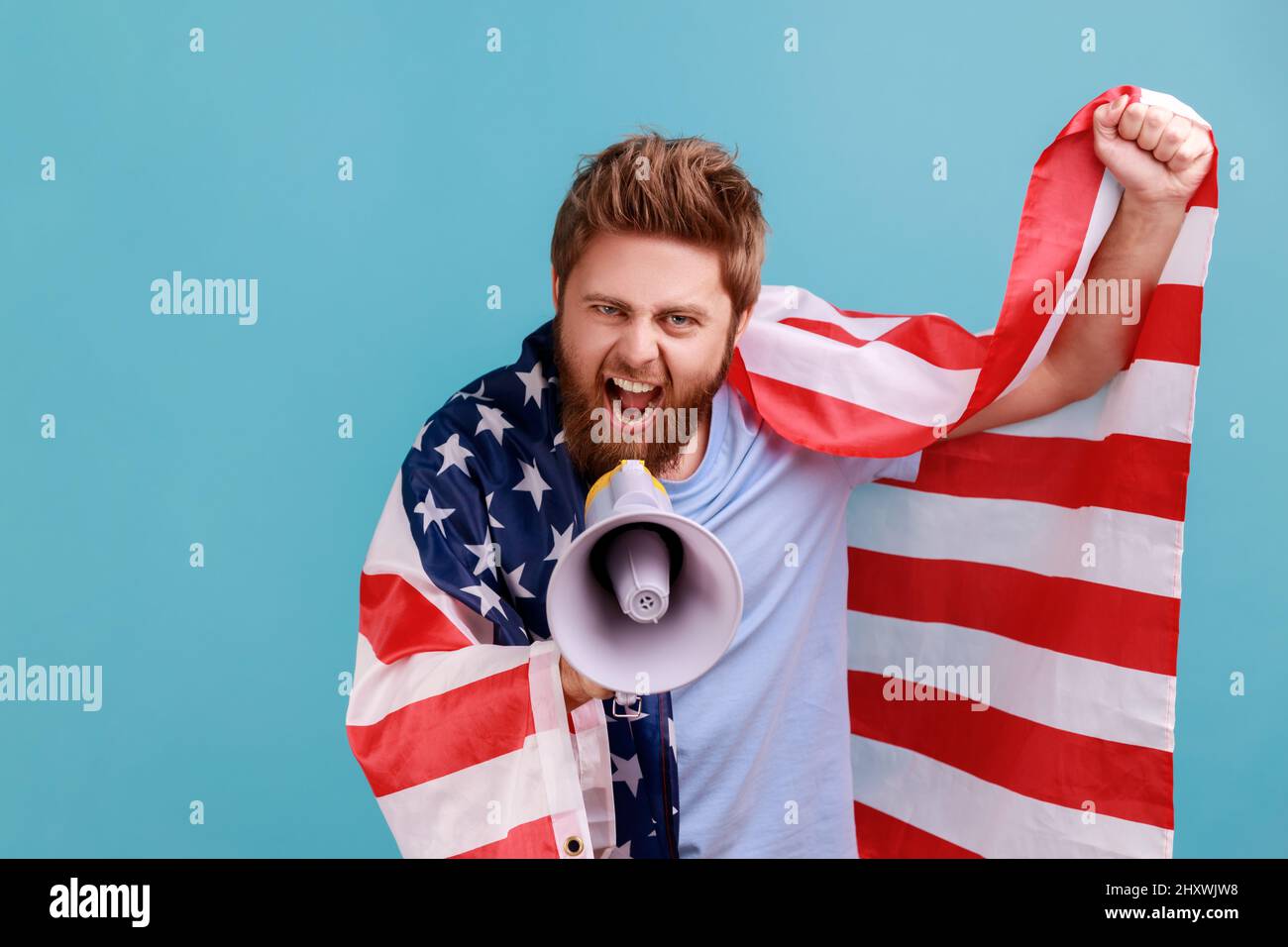 Portrait of excited self-confident bearded man screaming at loudspeaker holding flag of united states of america, patriotic protest. Indoor studio shot isolated on blue background. Stock Photo