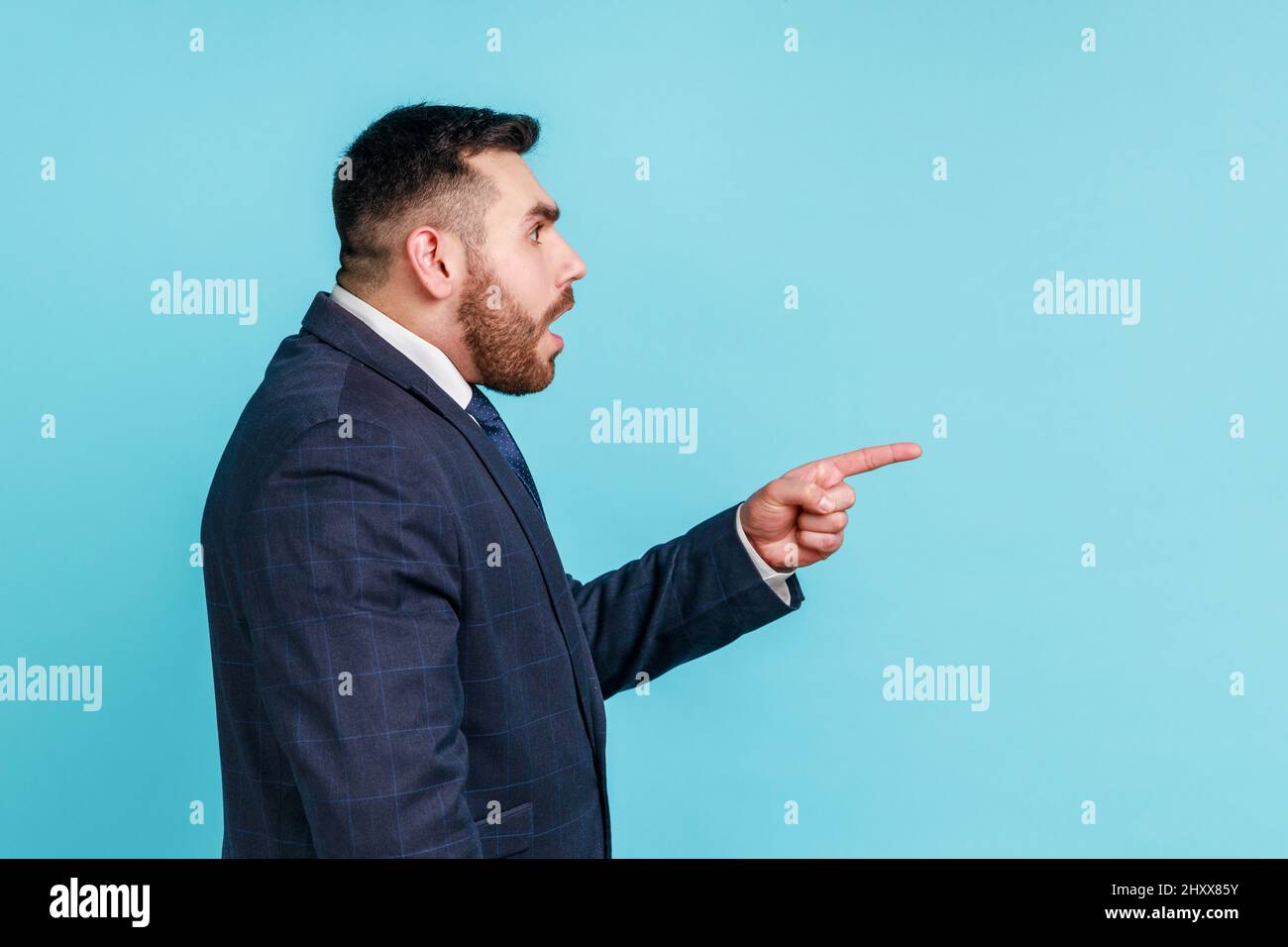 Portrait of attentive handsome bearded young man wearing official style suit having shocked facial expression, pointing finger aside. Indoor studio shot isolated on blue background. Stock Photo