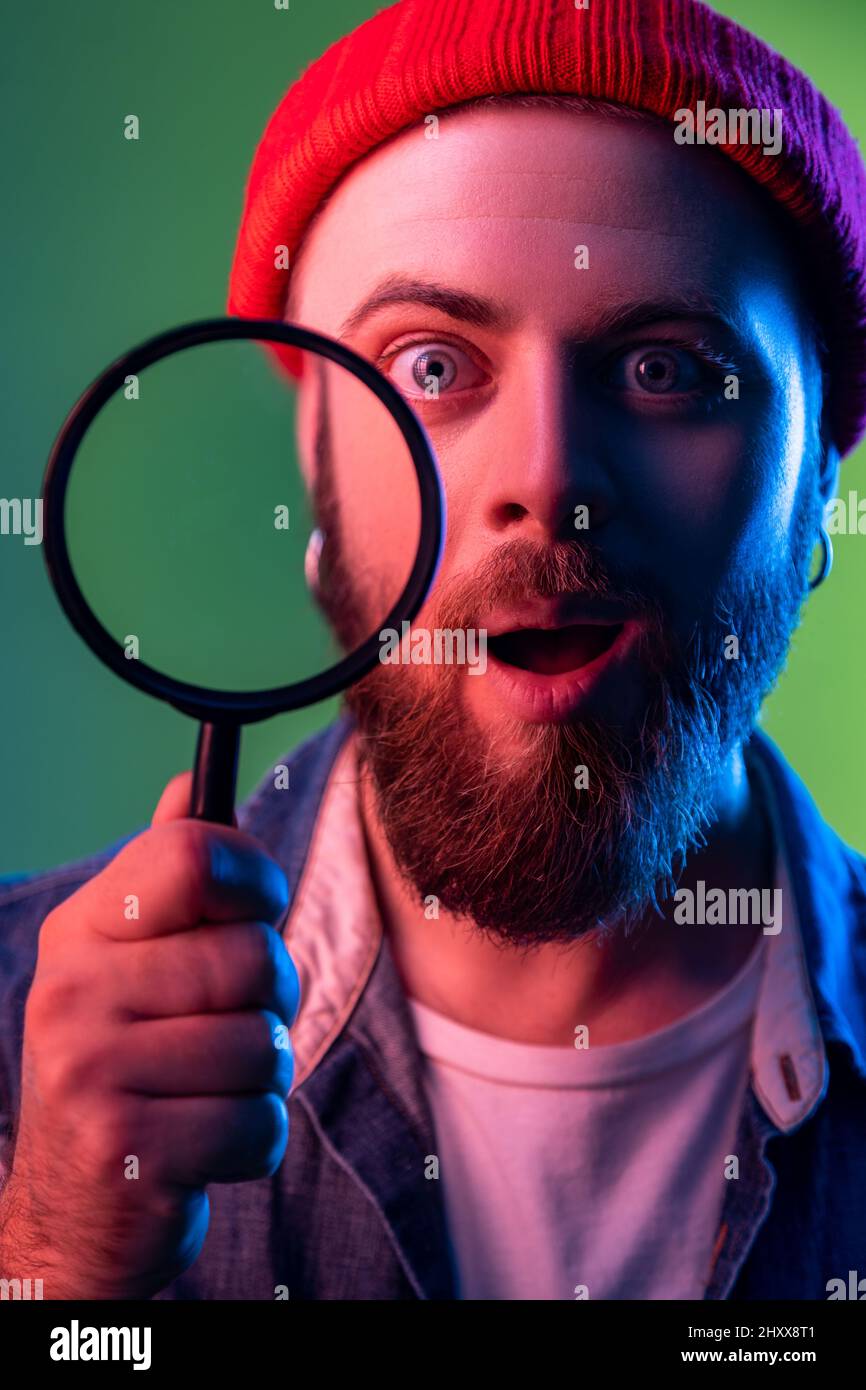 Portrait of amazed hipster man standing, holding magnifying glass and looking at camera, having astonished surprised face. Indoor studio shot isolated on colorful neon light background. Stock Photo