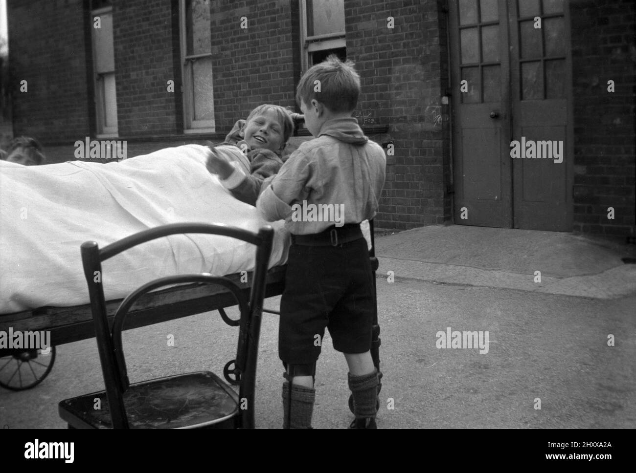 1942, historical, injured boy scout in leg braces, standing outside talking to another boy lying in a metal bed, Queen Mary's Hospital in Carshalton, Surrey, England, UK. Opened in 1908 as the Southern Hospital, in 1909 it became a place for the poor and sick children of London, many who had TB and was renamed the Children's Infirmary. The young patients were housed in single-storey ward blocks or 'cottages'. In 1915 it was renamed Queen's Mary Hospital for Children when Her Majesty became its patron. During WW2 the boy scouts who injured during the London Blitz were sent there for to recover. Stock Photo