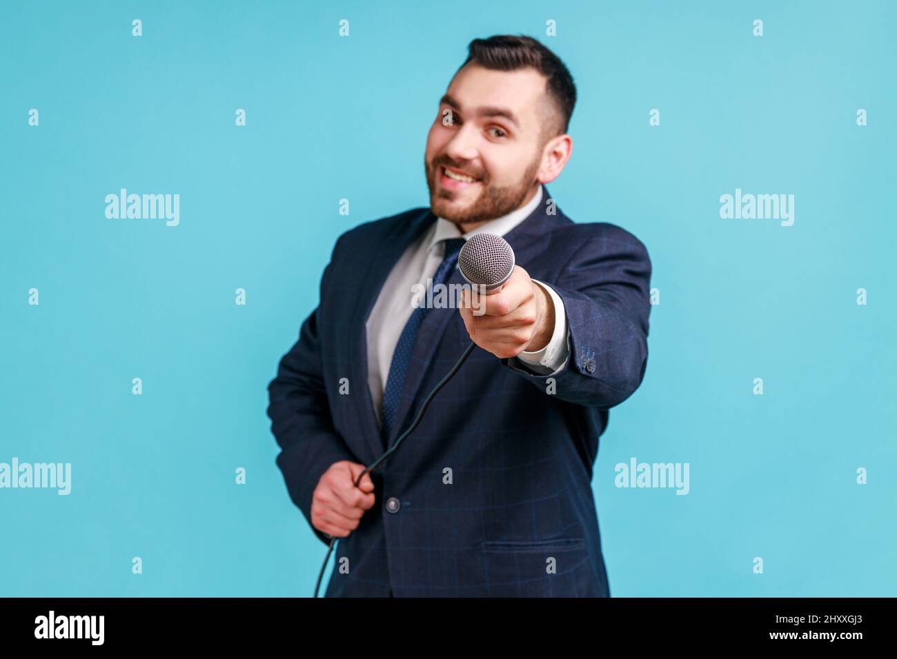 Smiling man journalist wearing official style suit holding microphone, making interview and asking opinion, looking at camera with smile. Indoor studio shot isolated on blue background. Stock Photo
