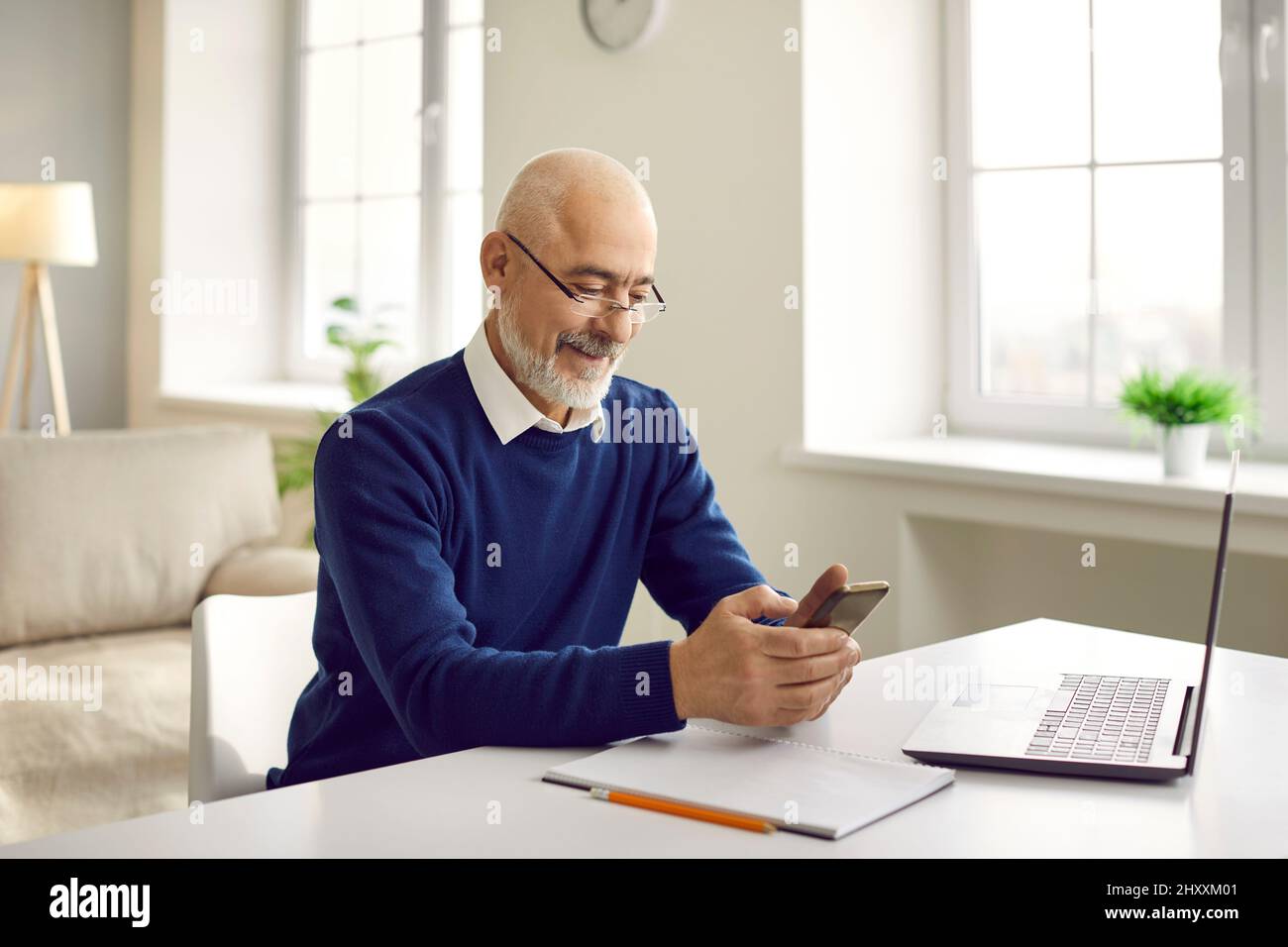 Happy senior man using his mobile phone while sitting at table with laptop computer Stock Photo