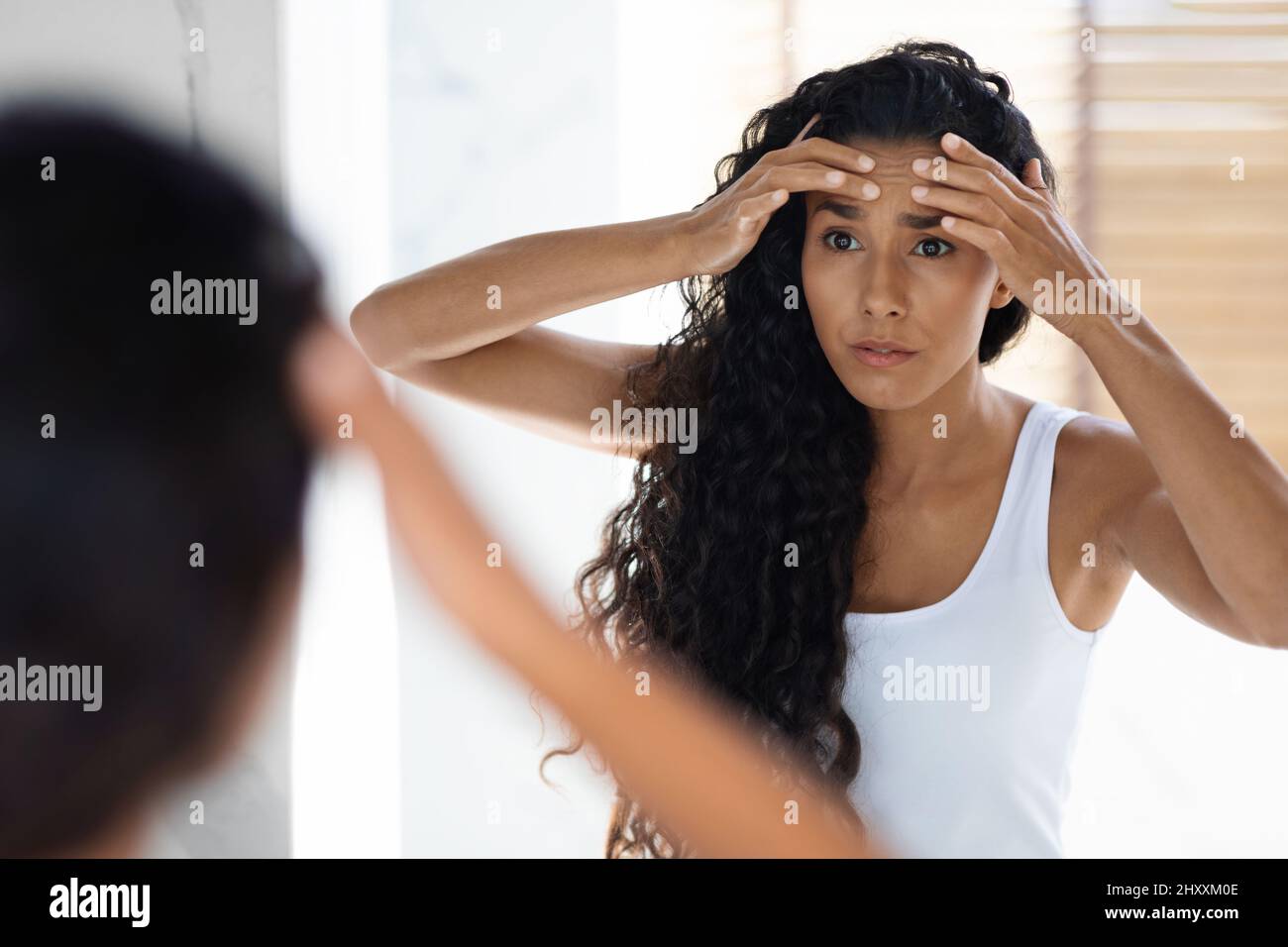 Worried Young Female Looking In Mirror And Touching Wrinkles On Her Face Stock Photo